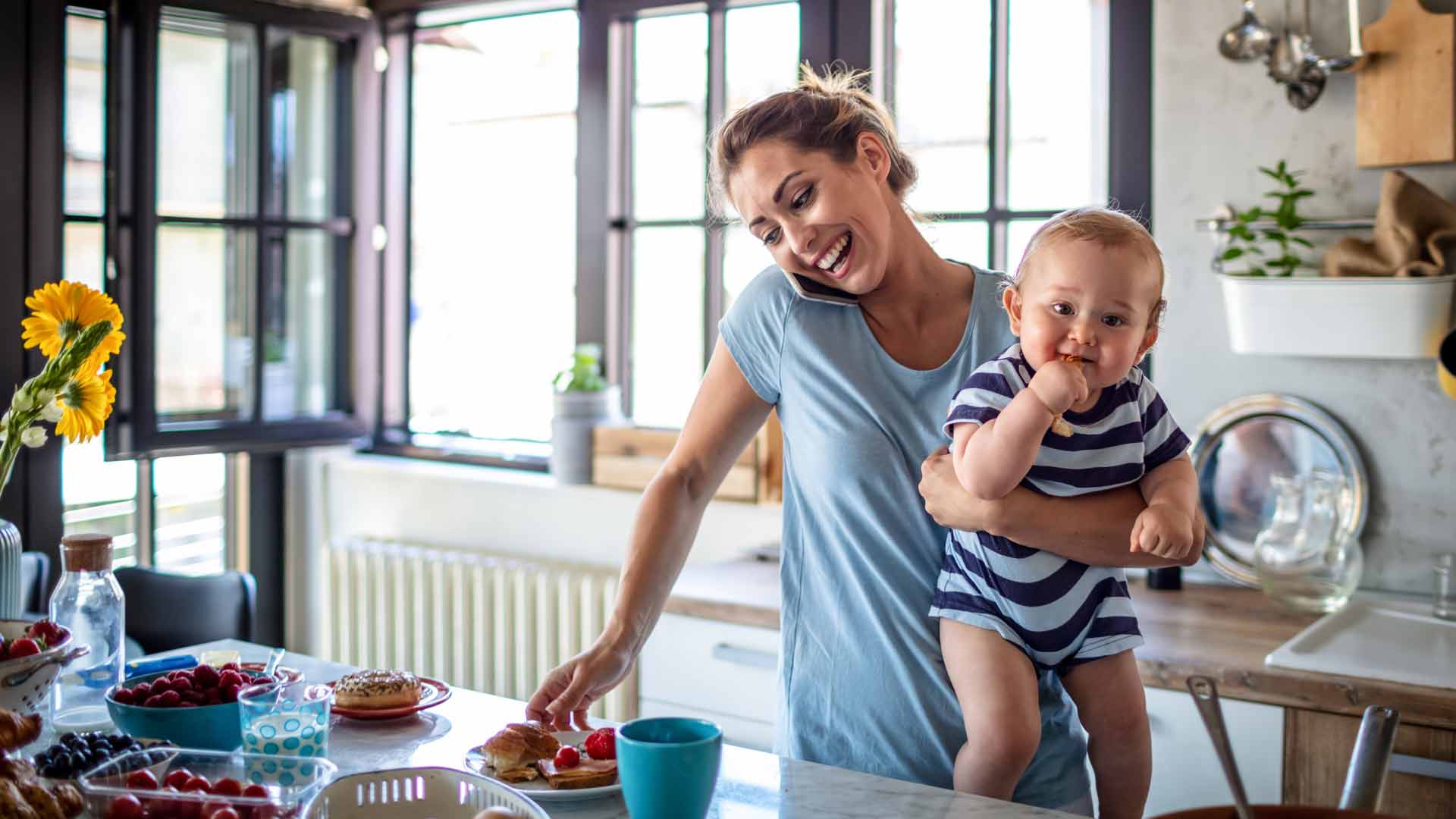 Frau mit Kleinkind auf dem Arm steht telefonierend in der Küche.