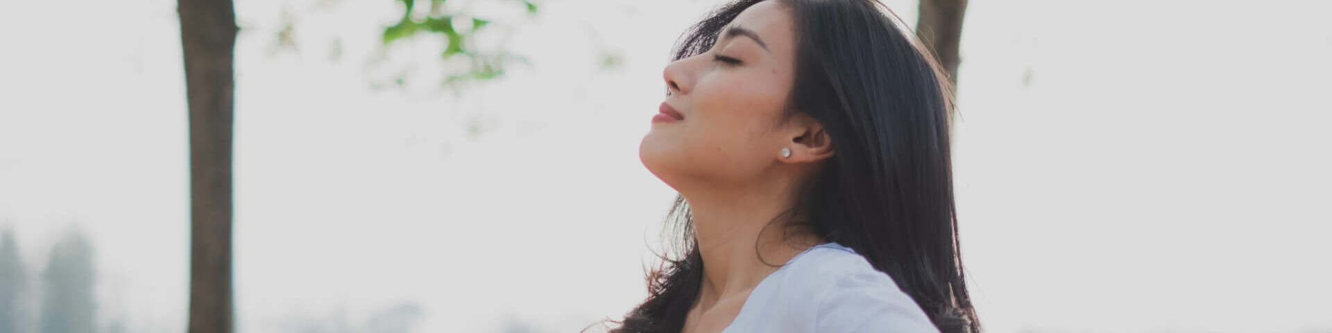 Frau mit dunklen Haaren und weißem T-Shirt atmet draußen frische Luft ein.