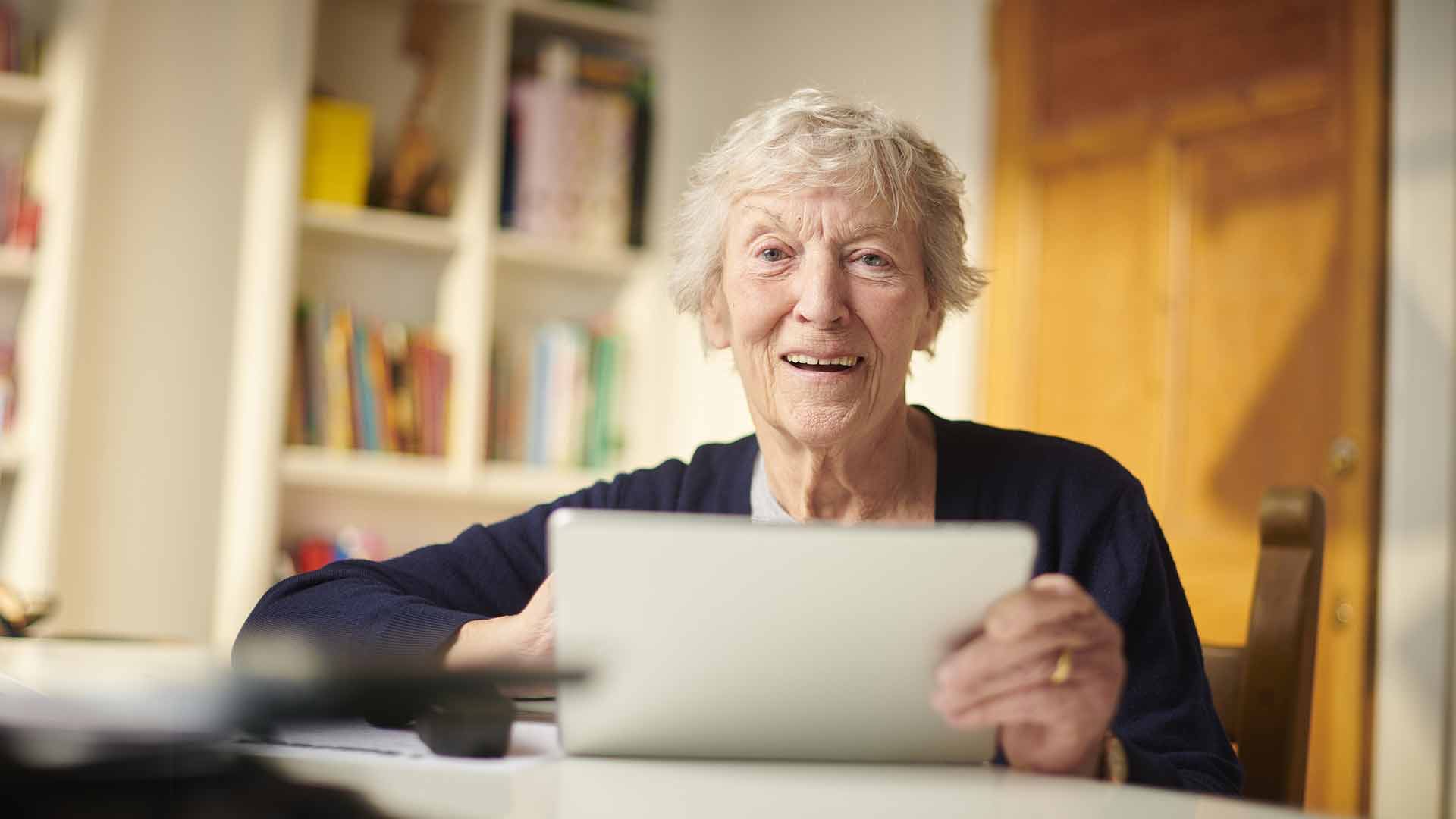 Ältere Frau mit kurzen Haaren hält in ihrem Zimmer ein Tablet in den Händen.