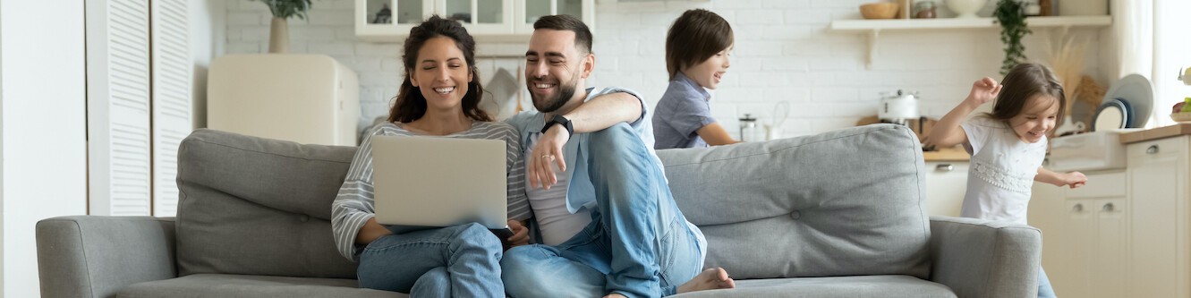 Ein Paar sitzt auf dem Sofa und schaut sich lächelnd auf dem Laptop etwas an, während zwei Kinder im Hintergrund fangen spielen.
